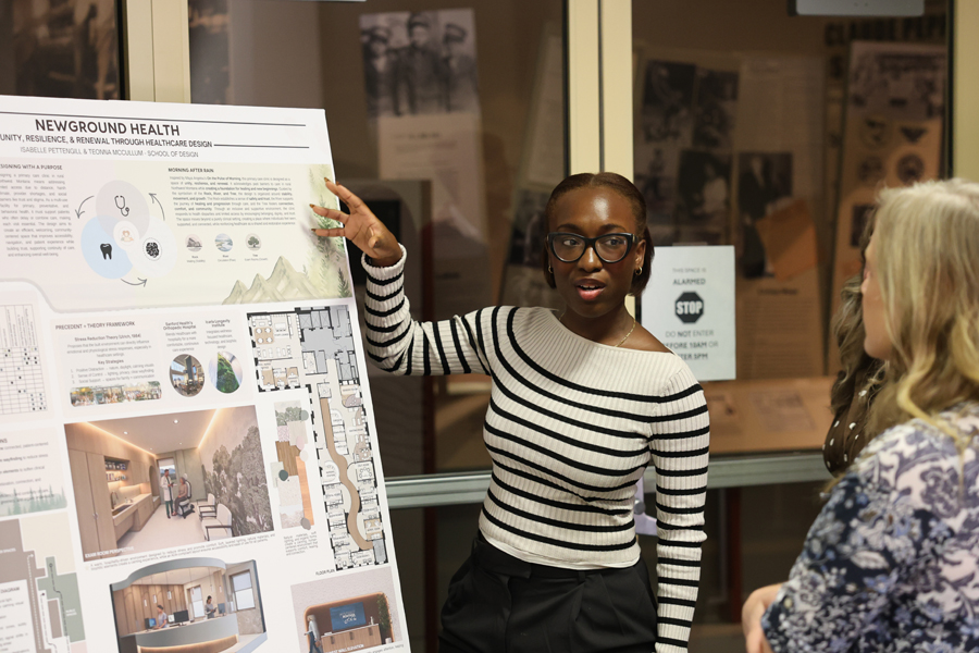 A presenter gestures while speaking with an attendee beside a healthcare design board and an