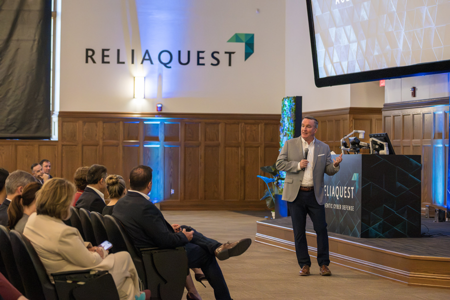 Mike McPherson speaking to a seated audience in an auditorium with a wood-beamed ceiling, with ReliaQuest logos on the walls and a large projection screen above the lectern.
