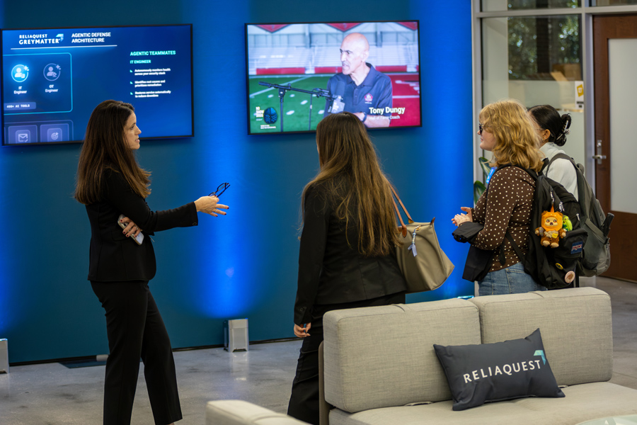 A ReliaQuest representative speaks with three students in a blue-lit indoor lounge featuring digital displays on ReliaQuest GreyMatter and a ReliaQuest-branded pillow on a sofa in the foreground.