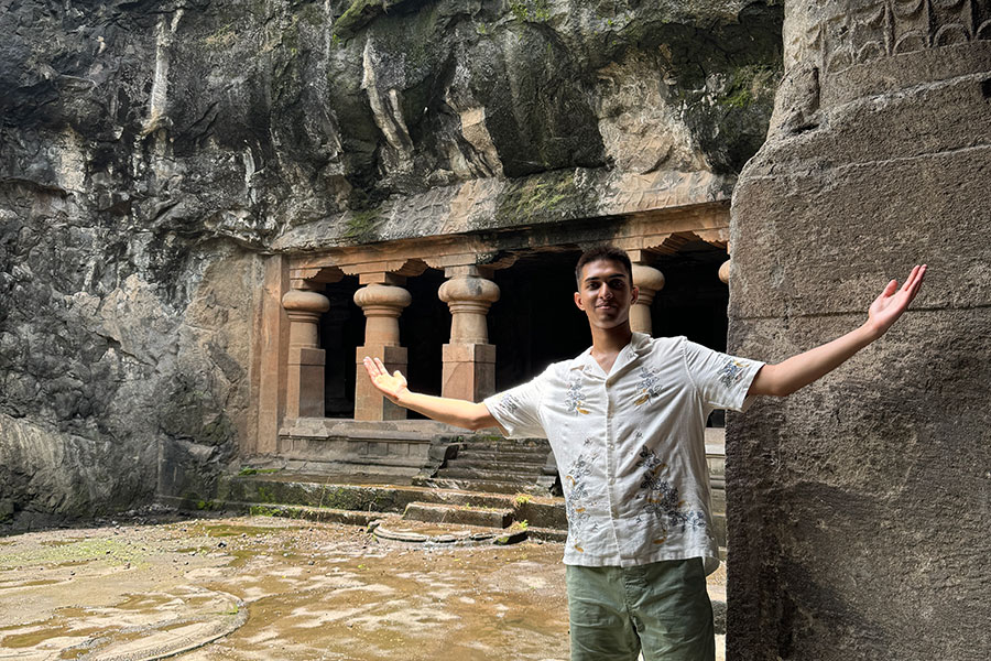 A young gentlemen poses with open arms in front of the Elephanta Caves in India.