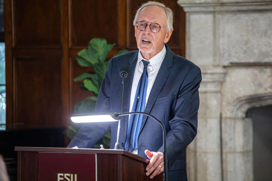 A person wearing a blue suit speaks while standing behind a dais. The FSU logo appears in the bottom right corner of the image.