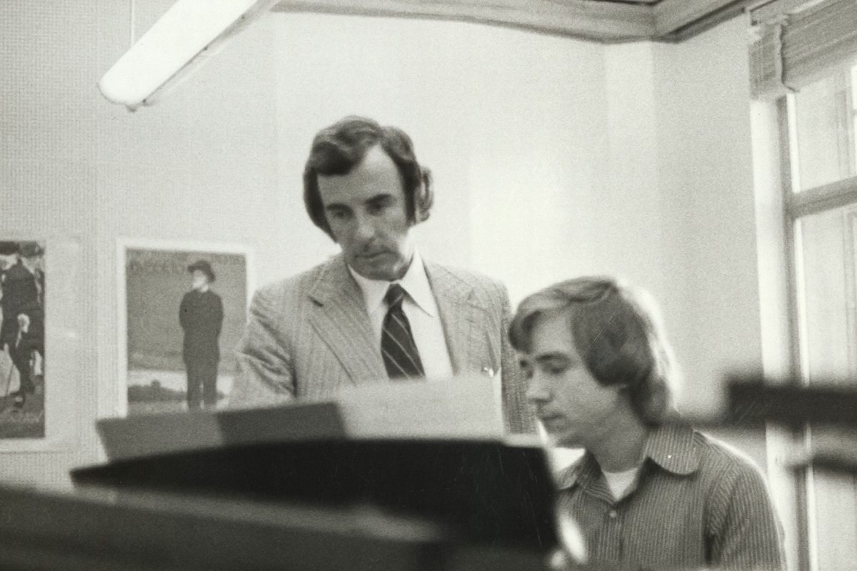 Black-and-white archival photograph of composer Carlisle Floyd standing beside a student seated at a piano, looking at sheet music during a music lesson.