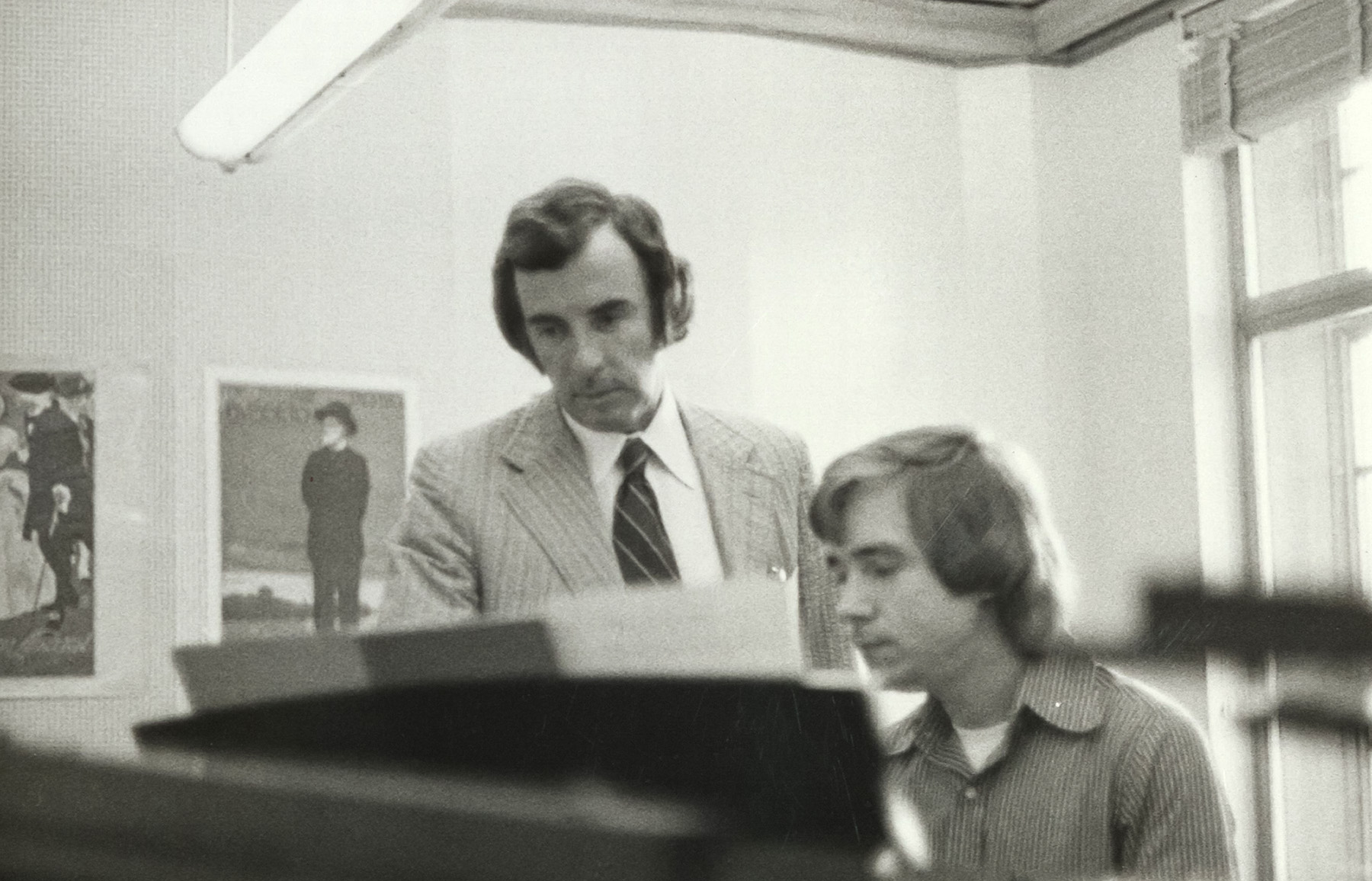 Black-and-white archival photograph of composer Carlisle Floyd standing beside a student seated at a piano, looking at sheet music during a music lesson.