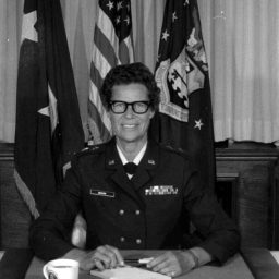 A black and white photo of Norma Elaine Brown in uniform smiling at a desk with flags behind her.