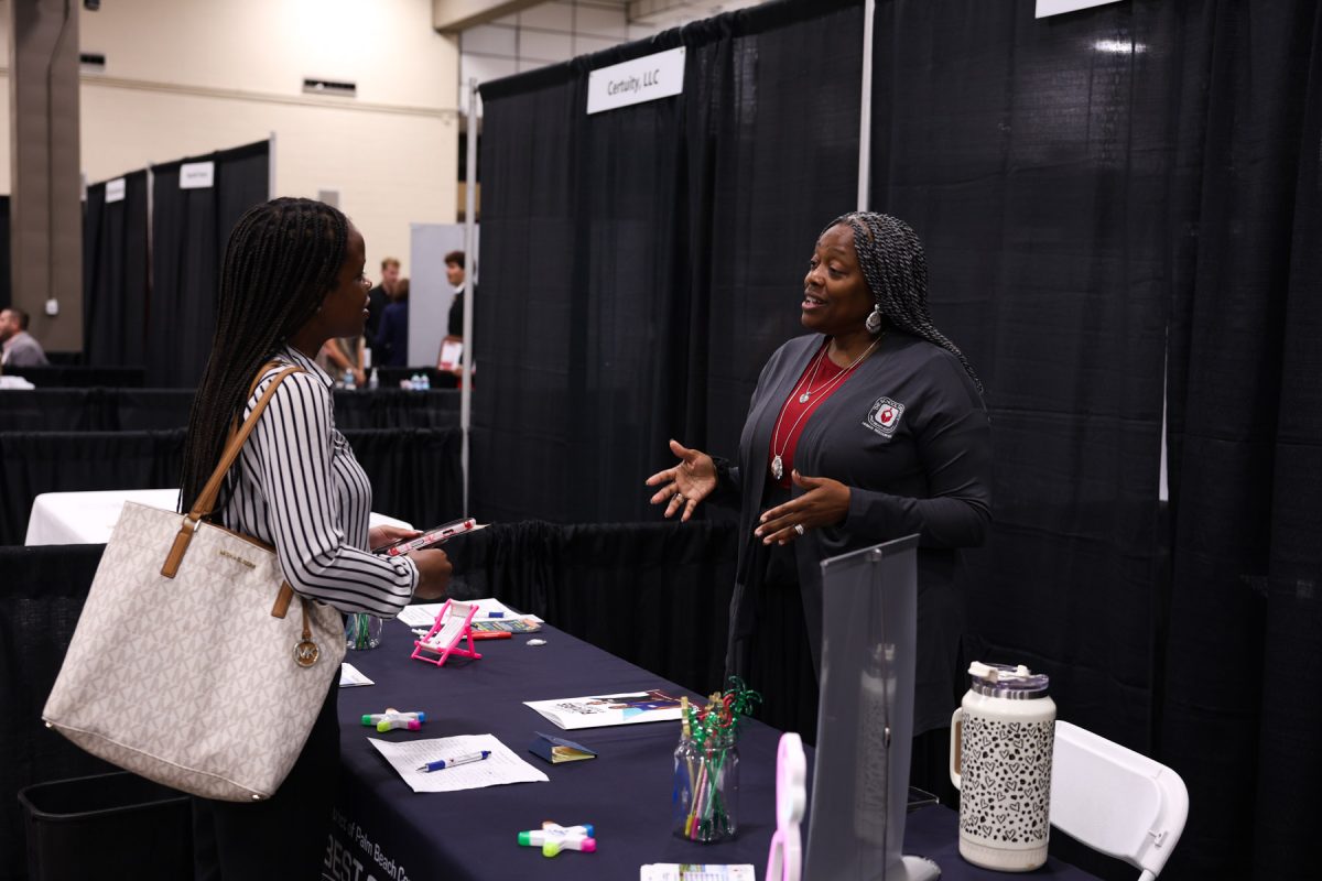 A student carrying a tote bag speaks with an employer representative behind a display booth at a career fair.
