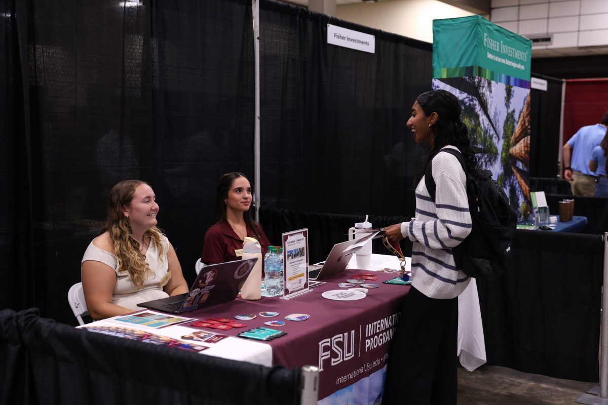 A student wearing a striped sweater and backpack smiles while speaking with two representatives seated at an FSU International Programs information table.