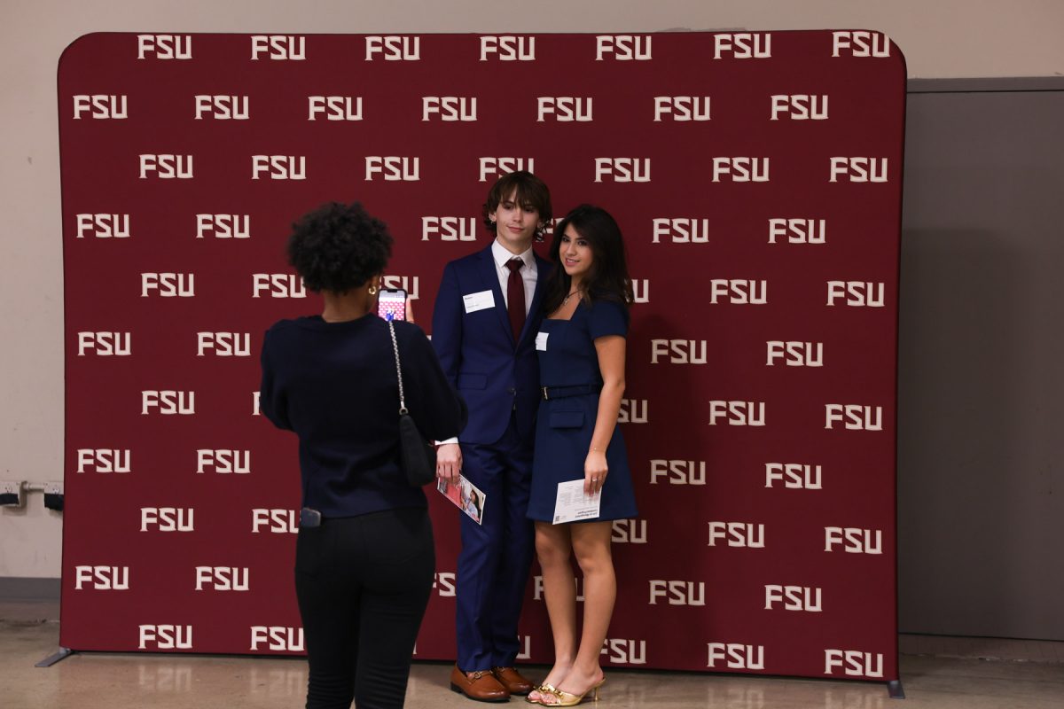 Two students in formal attire pose together in front of an FSU-branded backdrop while a third person takes their photo with a smartphone.