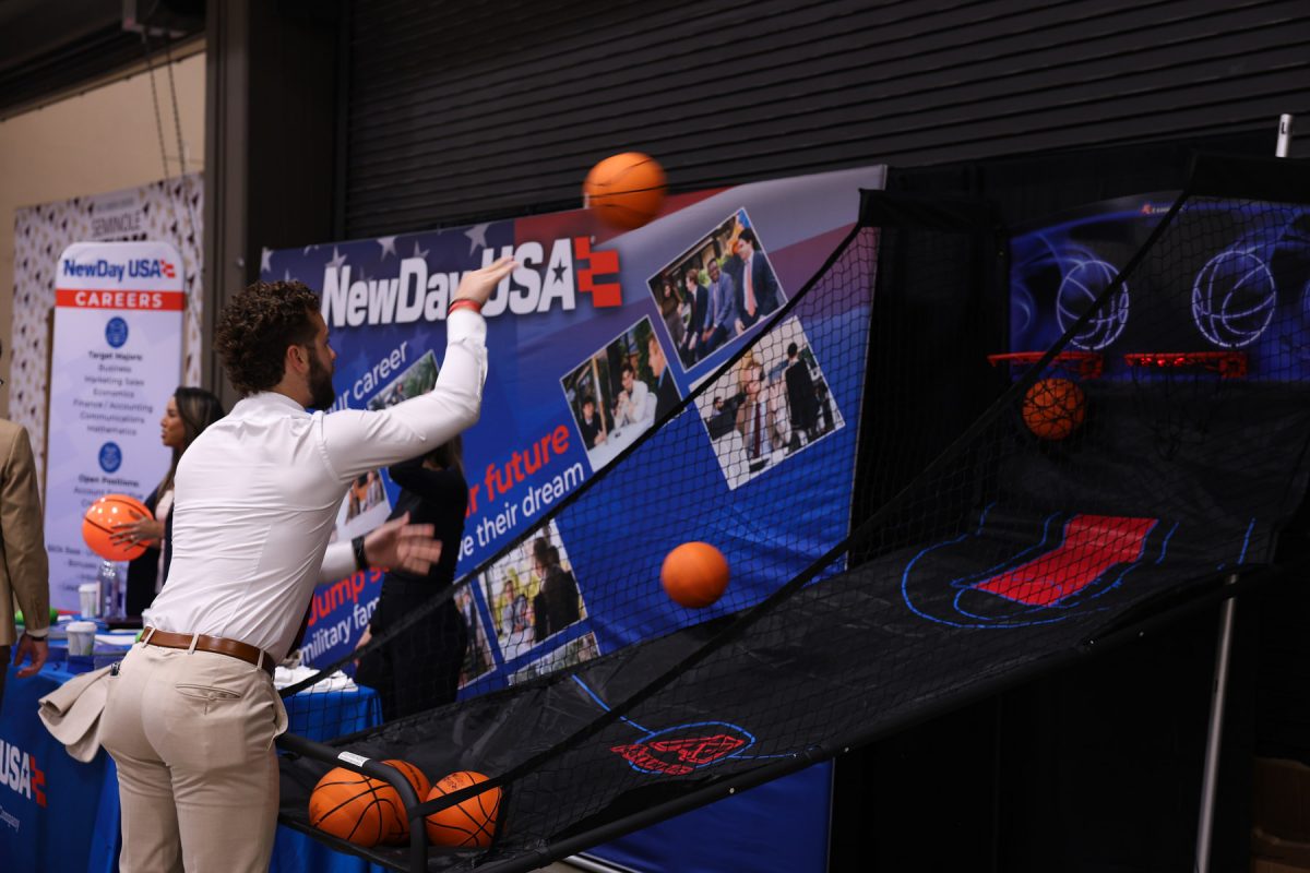 A student shoots a mini basketball at an arcade-style hoop set up in front of a company's promotional booth.