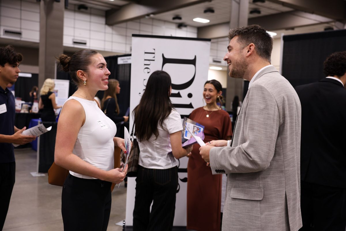 A student wearing a white top smiles while conversing with a recruiter in a light grey suit jacket at a busy career fair booth.