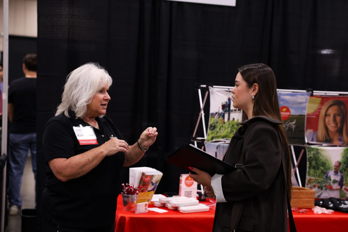 An employer representative gestures while speaking with a student holding a padfolio at a career fair booth draped in black.