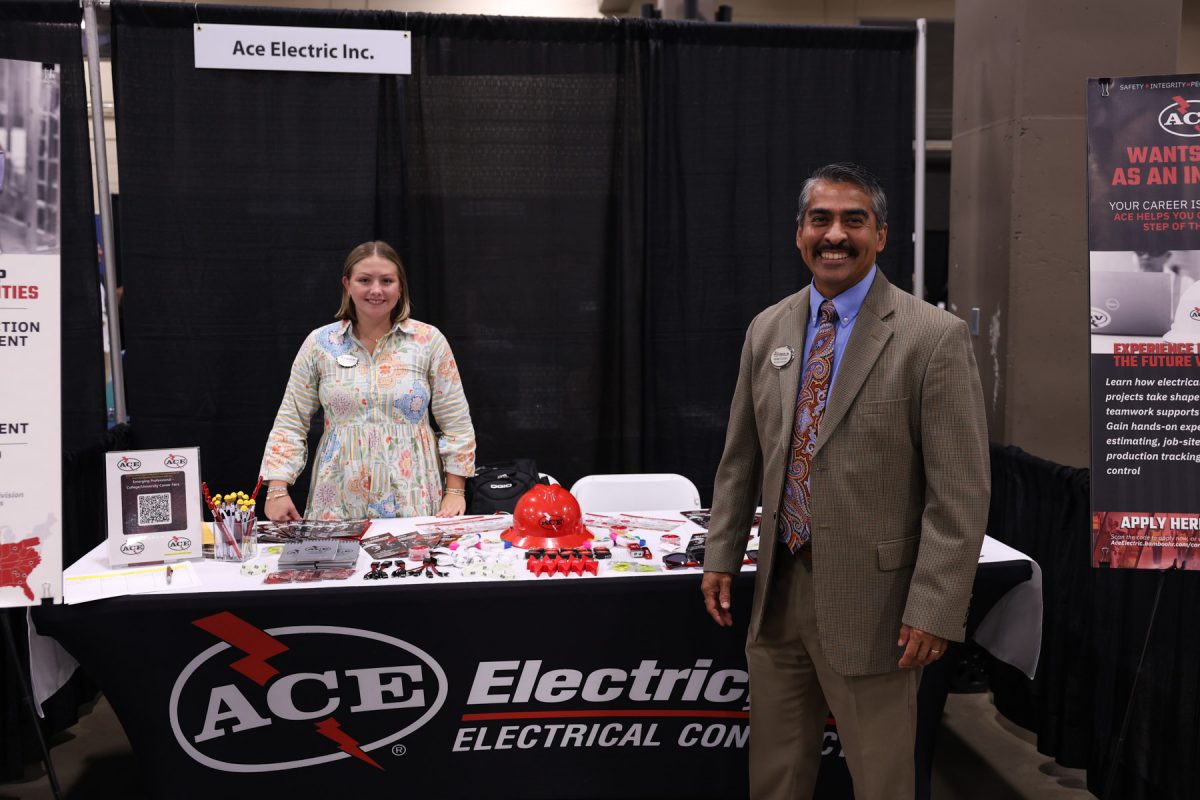 Two recruiters for Ace Electric Inc. smile behind and beside a company table displaying promotional items and hard hats.