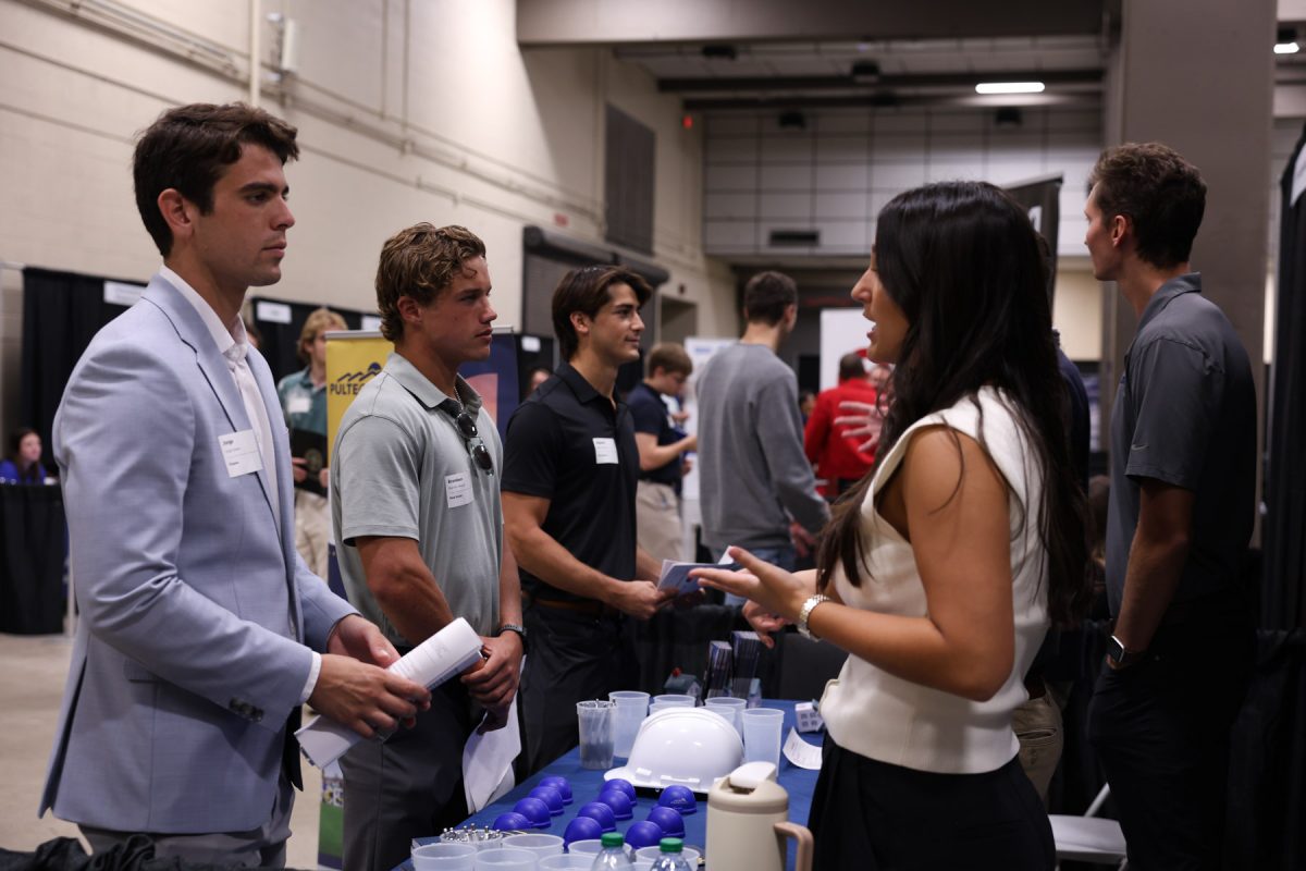 A group of students listens as a female employer representative speaks at a booth with mini hard hats on display.