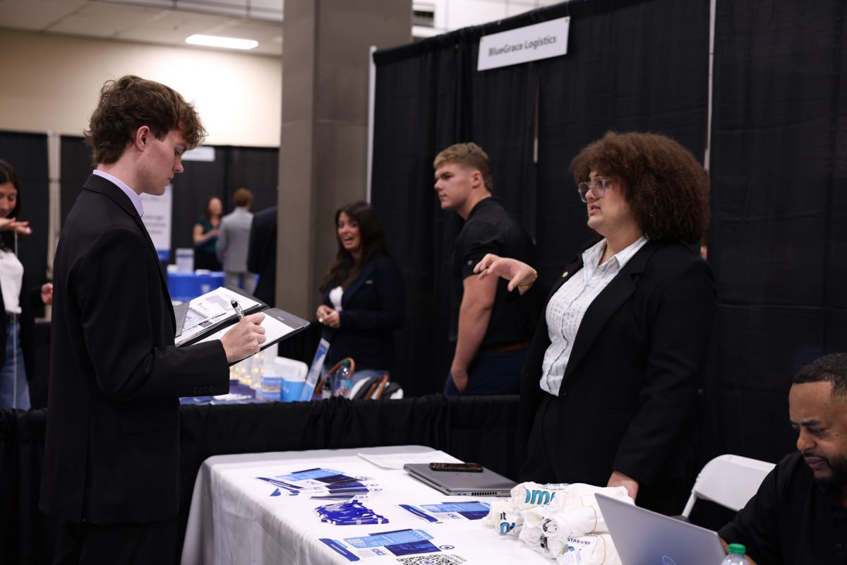 A student in a dark suit writes on a clipboard while speaking with a recruiter behind an information booth.