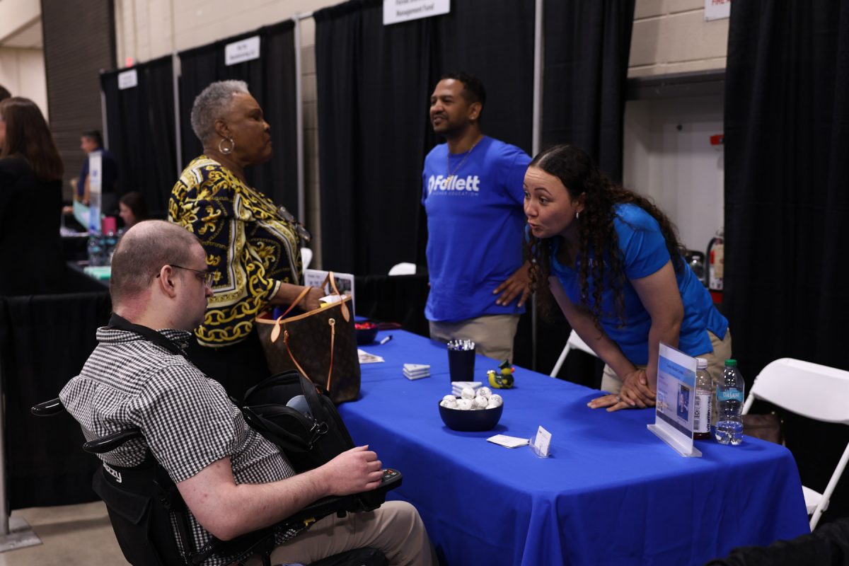 An employer representative leans across a blue table to speak with an attendee in a wheelchair at a career fair booth.