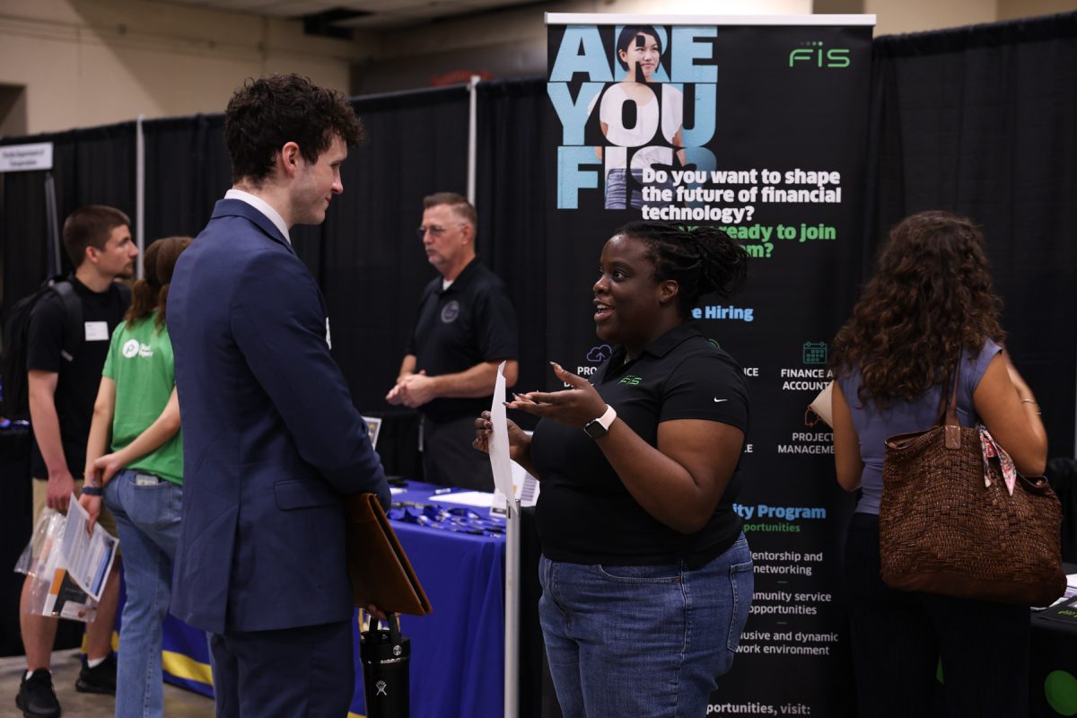 A recruiter in a black polo shirt speaks with a student in a blue suit holding a portfolio in front of an FIS company backdrop.