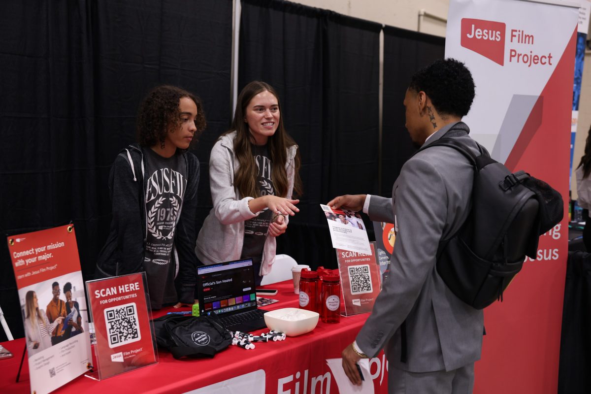 A student wearing a grey suit views a flyer while two representatives behind a red display table discuss the Jesus Film Project.