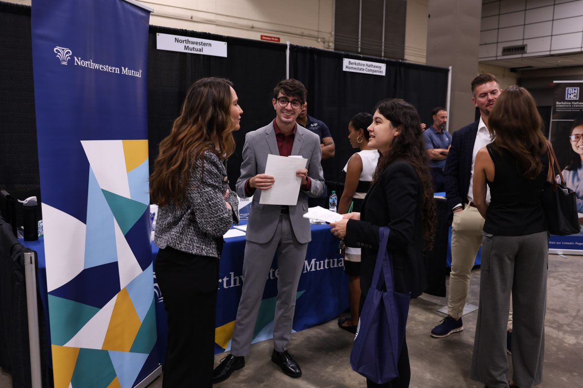 A male student holding papers smiles while conversing with a recruiter and another student in front of a Northwestern Mutual display.