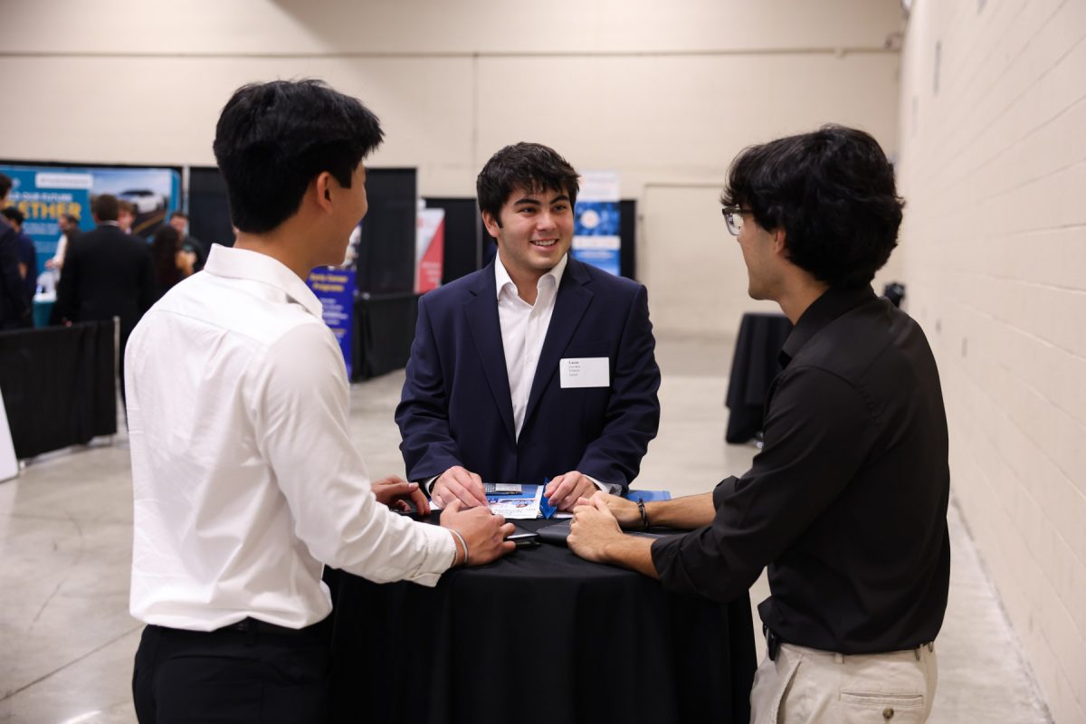 Three young men in business attire stand gathered around a tall, black table conversing during a career fair.