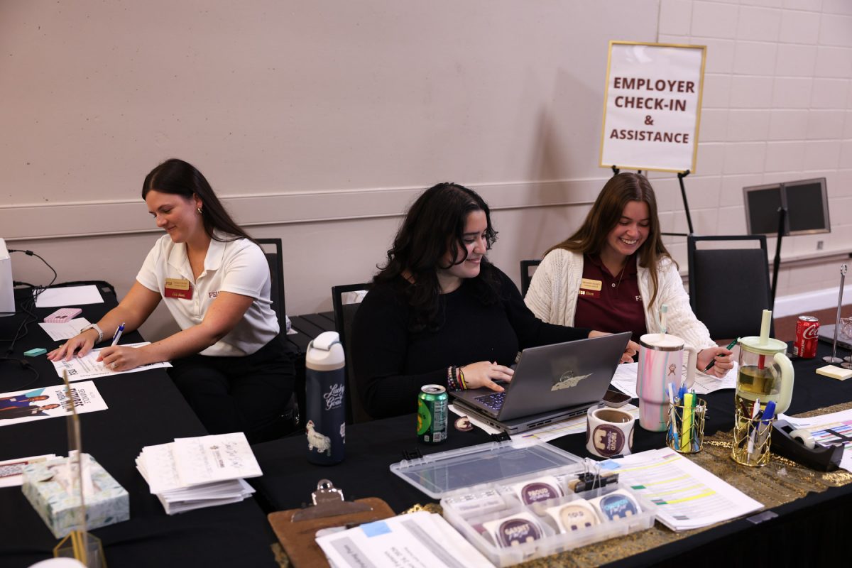 Three event staff members sit behind a registration table labeled "Employer Check-In & Assistance" working on laptops and paperwork.