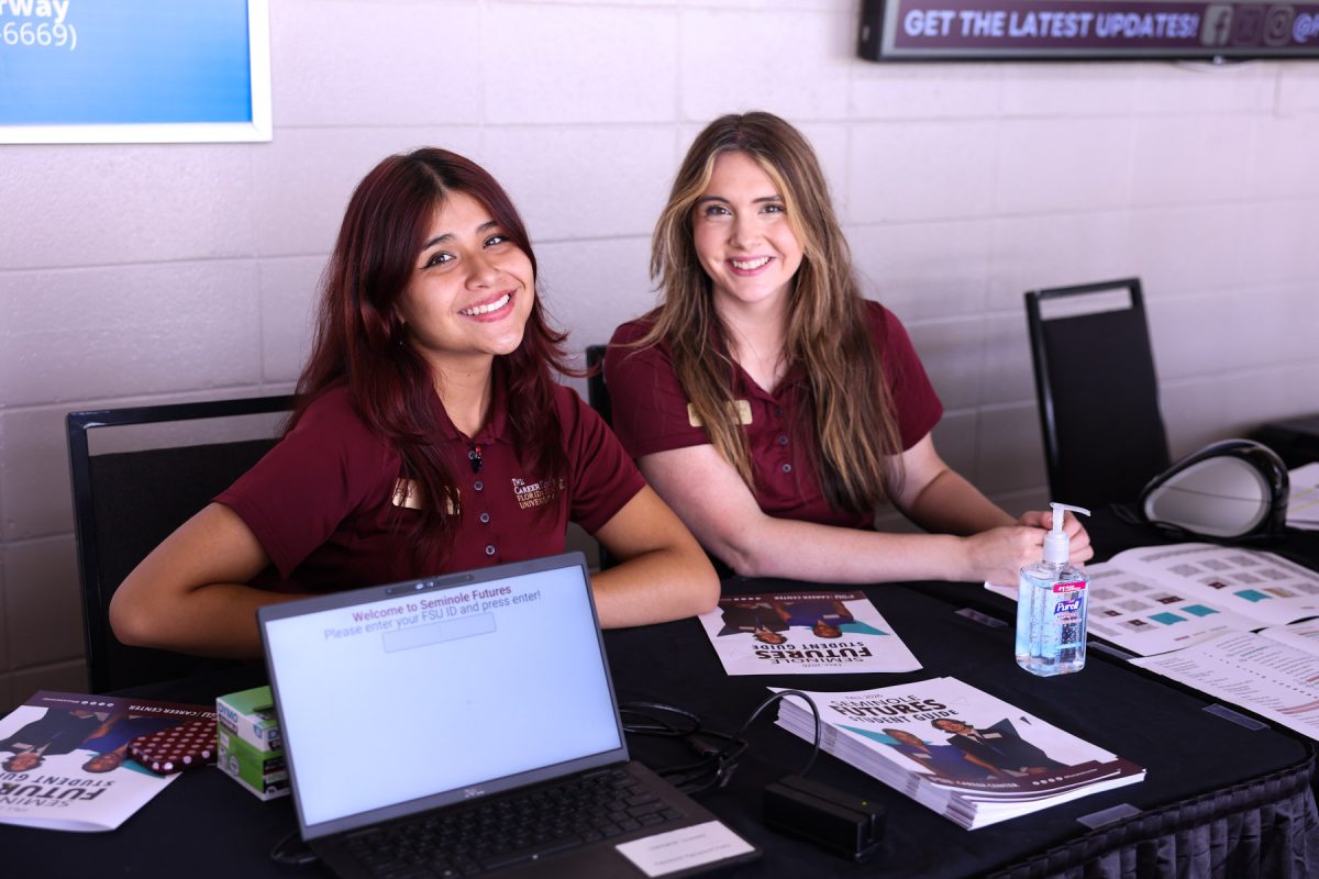 Two FSU Career Center student staff members wearing garnet polo shirts smile at a check-in table equipped with a laptop and event guides.